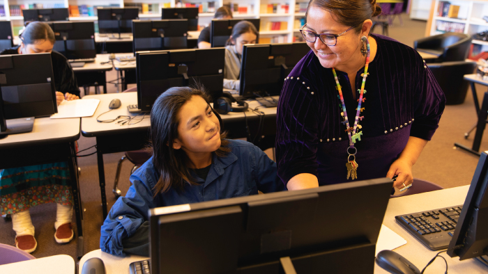 A smiling teacher assists a boy who is using a computer.