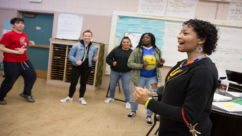 A female teacher with brown skin talks to a diverse group of students standing in a circle.