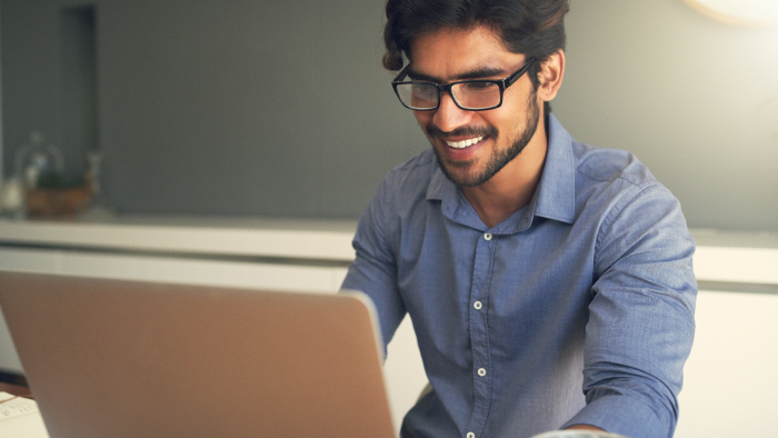 A male teacher with brown skin and glasses smiles as he works on his computer.