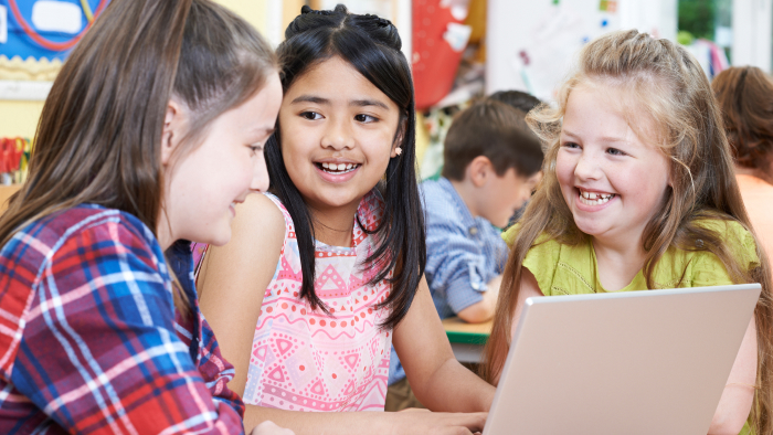 Three young girls interact while gathered around a laptop computer.