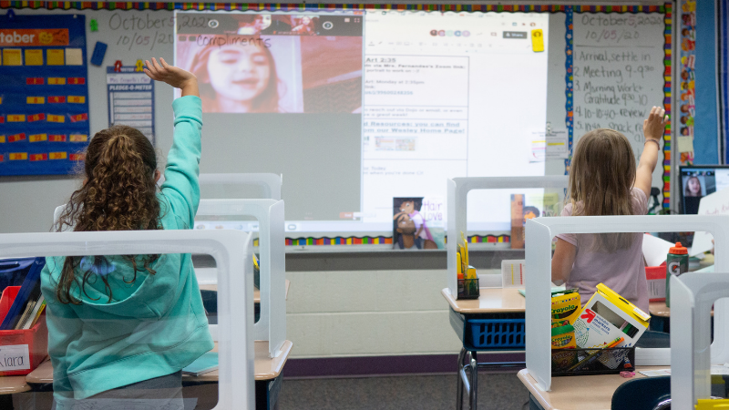 Students in a classroom wave to their online classmates