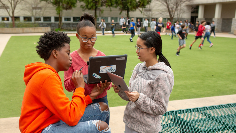 Photo of three students outdoors, looking at a laptop and talking