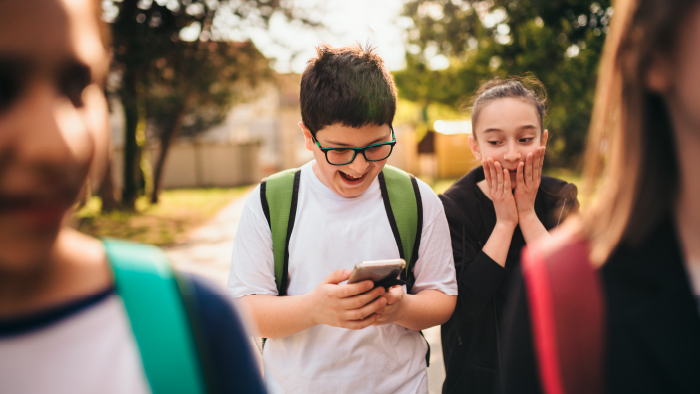 A boy looks at his phone, smiling, while a girl in the background covers her mouth in shock.