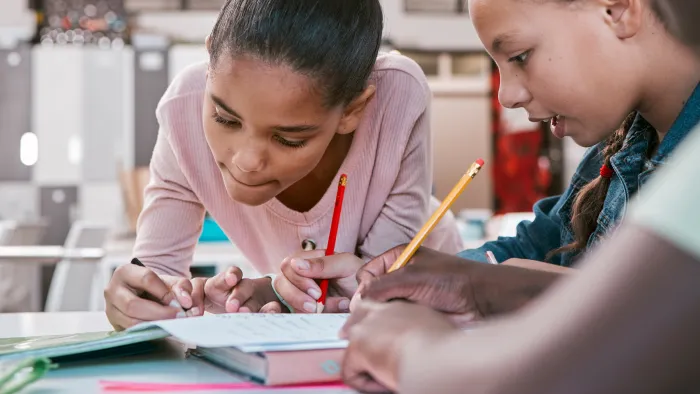 Three young students focus on the papers on their desks as they write or draw.